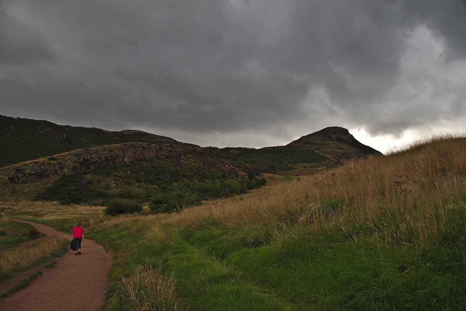 A walker on a red earth path through Holyrood Park with Arthur’s Seat under dark clouds ahead